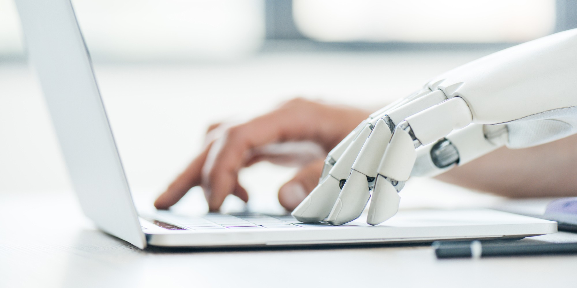 A robotic hand pairs with a human hand on a laptop to signify ai.
